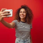 Young woman with curly hair taking a selfie against a red studio background with natural expression and soft editorial lighting.