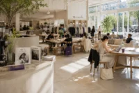 Bright fashion concept store with café seating, creative visitors, natural sunlight, and subtle violet accents in a modern editorial environment.