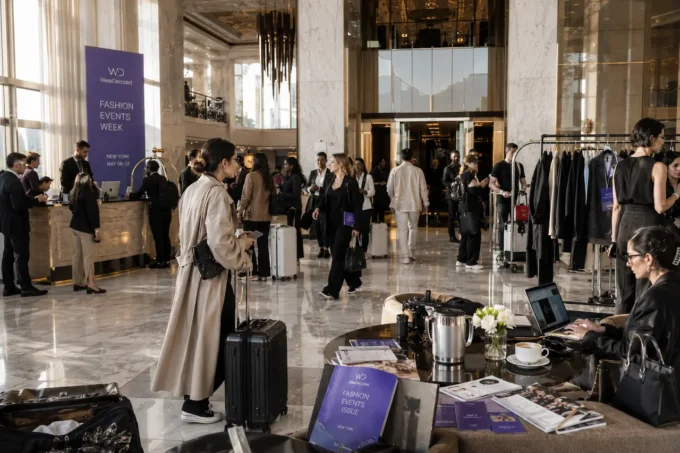 Fashion industry guests inside a luxury hotel lobby during event week with garment bags, stylish luggage, and bright editorial atmosphere.