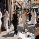 Women wearing relaxed minimalist dresses at a sunny café district in a modern city street-style editorial scene.