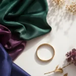Editorial fashion mood board with jewel-toned silk fabric, gold bangle, dried flowers, and soft natural daylight on a clean white background