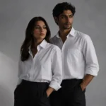 Editorial fashion portrait of a man and woman wearing oversized white shirts and black trousers in a minimalist studio with soft directional lighting and modern commercial styling.
