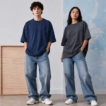 Two fashion models wearing oversized t-shirts and relaxed blue jeans posing inside a minimalist editorial studio with soft neutral lighting and modern streetwear styling.