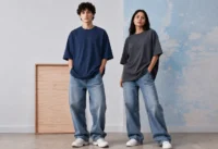 Two fashion models wearing oversized t-shirts and relaxed blue jeans posing inside a minimalist editorial studio with soft neutral lighting and modern streetwear styling.