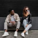 Ultra realistic street-style fashion portrait of a casually dressed young man and woman sitting against a black industrial wall, holding smartphones in a modern urban setting.