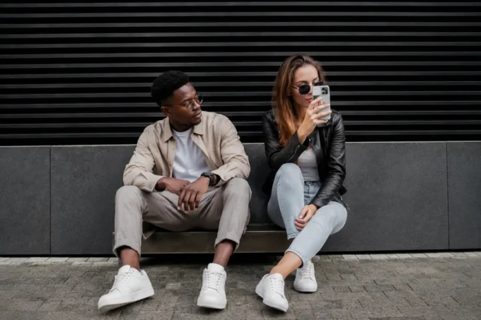 Ultra realistic street-style fashion portrait of a casually dressed young man and woman sitting against a black industrial wall, holding smartphones in a modern urban setting.