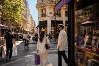 Stylish tourists walking through a luxury fashion shopping street filled with boutiques, cafés, and modern city atmosphere during daylight.